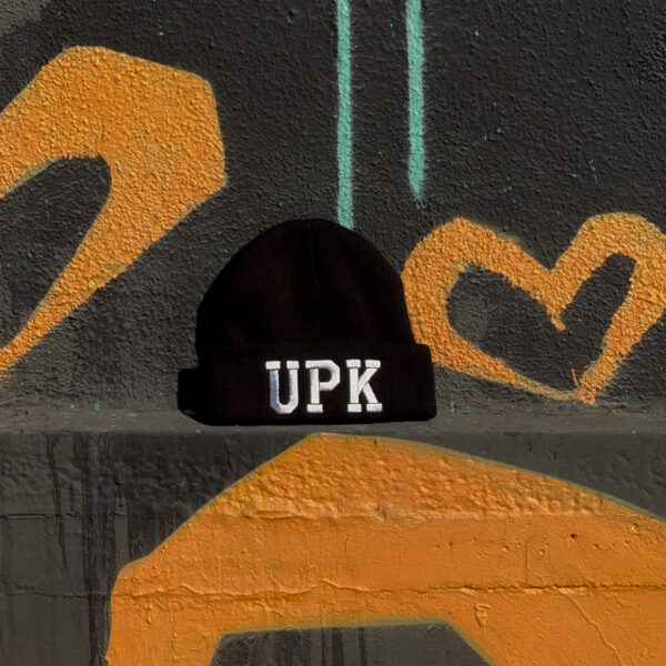 Black beanie with 'UPK' lettering against graffiti wall.