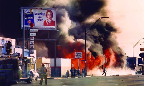 A large fire with thick black smoke engulfs a building while people watch from a distance.