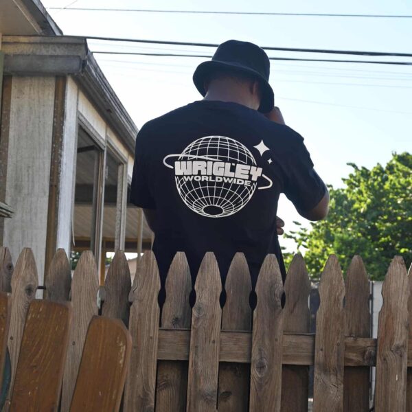 Person in a black hoodie with a graphic logo on the back, standing by a wooden fence.