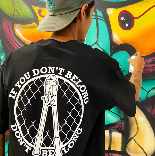Person in a cap painting a colorful mural, wearing a black shirt with a bold graphic and text.