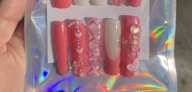 Close-up of intricately designed pink and red acrylic nails with rhinestones and heart shapes.